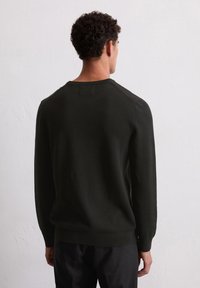 Homme aux cheveux courts et bouclés, portant un pull long noir à manches longues et un pantalon noir, faisant face à un mur blanc texturé.