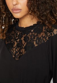 Μαύρο φόρεμα με λευκή δαντέλα στον γιακά, ρυτιδωμένο λαιμό και λεία υφή. Ο σχεδιασμός συνδυάζει διαφανείς και αδιαφανείς υφές.