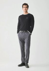 Bărbat stand cu mâinile în buzunare, purtând un pulover negru, pantaloni gri, curea neagră și pantofi negri, pe un fundal alb simplu.