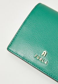 Roheline tekstureeritud nahkrahakott nähtavate õmblustega ja hõbedane Furla logo valgel taustal.