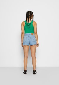 Grünes ärmelloses Oberteil, hellblaue Jeansshorts mit fransigem Saum und schwarze Sneakers. Die Shorts verfügen über Taschenverzierungen und ein geprägtes Leder-Patch.