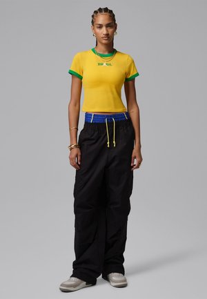 Kvinde med fletninger iført en gul Brasilien T-shirt, sorte vide bukser med blåt bånd, hvide sneakers og guldsmykker, stående mod en grå baggrund.