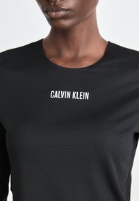 Close-up van een persoon die een zwart gestructureerd shirt draagt met erop in het midden van de borst de witte tekst "CALVIN KLEIN".