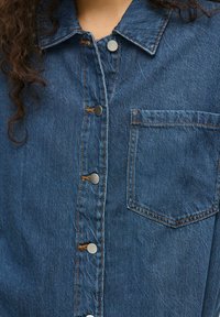 Camicia di denim con bottoni a pressione argentati e una tasca sul petto, indossata da una persona con capelli ricci e scuri.