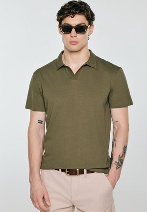 Mężczyzna noszący oliwkowo-zielone polo z krótkim rękawem, beżowe spodnie, ciemne okulary przeciwsłoneczne i pasek, z tatuażami na obu przedramionach, stoi na jednolitym tle.