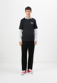 Young man standing against white wall wearing black "Empire" T-shirt over striped shirt, black pants, and red sneakers with white soles.