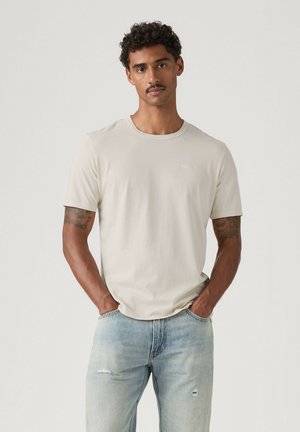 Uomo in piedi con le mani in tasca che indossa una maglietta beige chiaro e jeans azzurri strappati su uno sfondo semplice.