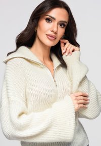 Pull en maille épaisse de couleur crème avec un col haut et une fermeture éclair. Caractérisé par des manches larges et un motif texturé sur l'ensemble.
