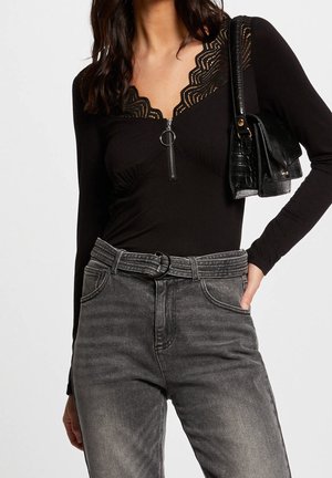 Kvinde iført sort lynlåstop med blonder, højttaljede grå jeans med bælte og bærende en sort skuldertaske.