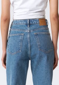 Οπίσθια όψη ατόμου που φοράει ψηλόμεσα μπλε τζιν με ραμμένες τσέπες και καφέ δερμάτινο ταμπελάκι με την ένδειξη «WEEKDAY».