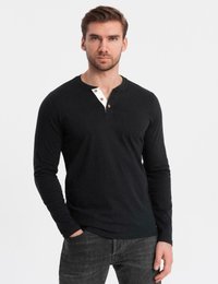 Man i svart långärmad henleytröja med vit knappplacket och mörkgrå jeans, stående mot vit bakgrund.