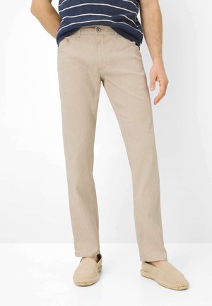 Mand iført beige bukser med lige ben, en stribet marineblå og hvid skjorte samt beige slip-on sko, der står foran en hvid baggrund.