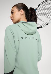 Lys grønn hettegenser med teksturert stoff, som har en svart "ENDLESS" logo på baksiden og hette. En racket holdes over skulderen.