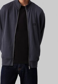 Man som bär en mörkgrå dragkedjajacka över en svart skjorta och mörkblå jeans, stående mot en slät ljusgrå bakgrund.