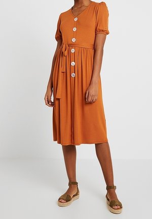 Vrouw draagt een roestoranje jurk met knopen aan de voorkant, tot op de knieën, met korte pofmouwen en een taillekoord, gecombineerd met olijfgroene sandalen met enkelriem.