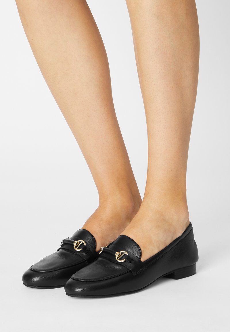 Mocassins en cuir noir avec détail de logo en métal doré portés sur jambes nues sur un fond blanc uni.