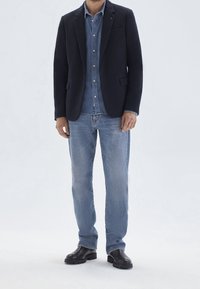 Man wearing a black blazer, blue denim shirt, light blue jeans, and black leather boots standing against a plain light background.