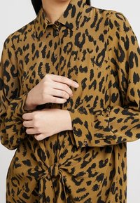 Blusa in stampa leopardata marrone con colletto, chiusura frontale a bottoni e maniche lunghe, caratterizzata da un dettaglio annodato in vita. Tessuto di superficie liscia.