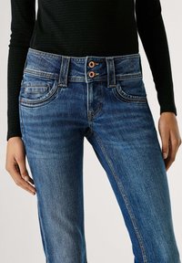 Džinsa bikses ar vidēji zilu mazgājumu, piecu kabatu dizainu, divu pogu aizdari, kontrastējošu šuvju apdari un viegli šaurinātām kājām.