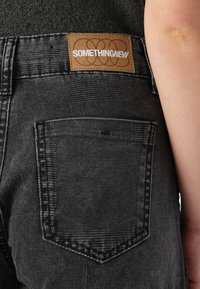 Črne jeans hlače iz denima z teksturiranim zaključkom. Imajo tanek usnjen etiket z napisom "SOMETHINGNEW" in zadnji žep z kontrastnim šivanjem.