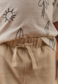 Smėlinės spalvos medvilniniai šortai su elastine juosta ir raišteliu, derinami su šviesios spalvos marškinėliais su saulės ir palmės motyvais.