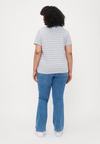 Camiseta de rayas con mangas cortas en azul claro, blanco y un suave tono marrón. Combinada con unos jeans acampanados azules y zapatillas blancas, vista desde la parte de atrás.