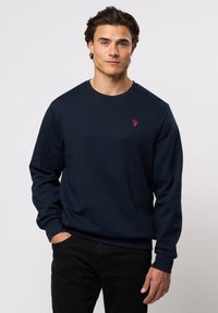 Mørkeblå hettegenser laget av bomull, med lange ermer, rund hals, og en liten rød logo på brystet. Svart bukse synlig.