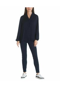 Navyblauwe blouse met lange mouwen en V-hals, gecombineerd met strakke navy pants en witte sneakers met zwarte accenten. Glad materiaal, ontspannen pasvorm.