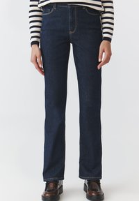 Jeans dritti in denim blu scuro a vita alta, con cuciture arancioni a contrasto e design classico a cinque tasche. Indossati con una maglietta a righe.