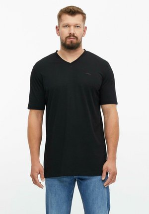 Mann mit kurzen Haaren und Bart, der ein schlichtes schwarzes V-Neck-T-Shirt und blaue Jeans trägt, blickt nach vorne vor weißem Hintergrund.