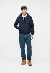 Νεαρός άνδρας με μπλε ναυτικό μπουφάν, λευκό πουκάμισο, μπλε τζιν με μεγάλες μπροστινές τσέπες και καφέ μποτάκια με κορδόνια, στέκεται μπροστά σε απλή λευκή φόντο.