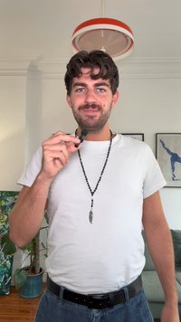 Um homem com uma t-shirt branca segura um microfone preto. Ele usa um colar longo com contas e um pendente de pena. O fundo apresenta uma planta.