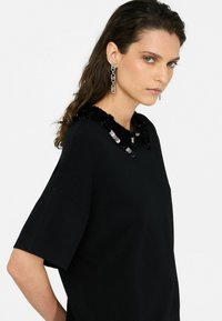 Black top with short sleeves and a textured, sequined collar. Model wears silver drop earrings, showcasing a simple yet elegant design.