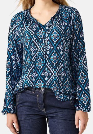 Blaue Bluse mit geometrischem Muster in Türkis und Weiß, mit einem gerafften Rundhalsausschnitt und langen Ärmeln. Mit dunklen Jeans kombiniert.
