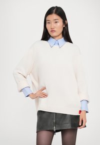Donna in un maglione bianco oversize sopra una camicia blu chiaro e una gonna di pelle nera con zip frontale, in posa con una mano sulla coscia contro uno sfondo bianco.