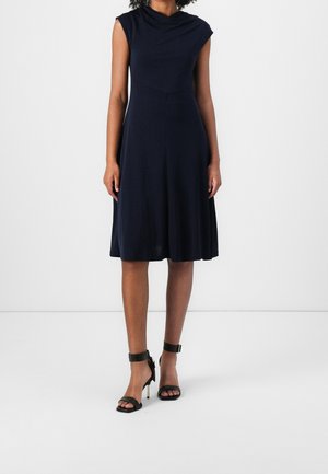 Mujer con vestido azul marino sin mangas hasta la rodilla y tacones altos negros con correa en el tobillo, de pie frente a un fondo blanco liso.