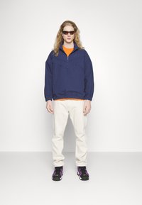 Jūras spēku vējjakas ar puszipu un elastīgu jostasvietu, oranža T-kreklu, krēmveida bikses un melnus sporta apavus ar purpura akcentiem.