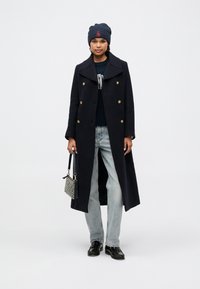 Marineblå uldfrakke med guldknapper, parret med en strikket marineblå hue, sort sweater, lyseblå jeans og blanke sorte loafers, bærende en mønsteret taske.