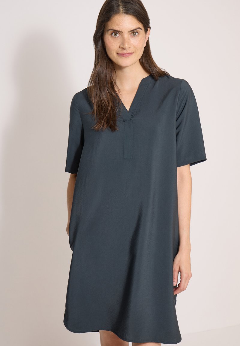 Femme aux longs cheveux bruns debout devant un fond uni, portant une robe gris foncé à manches courtes, à col en V et coupe ample.