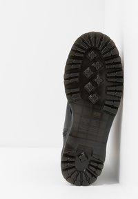 Semelle de botte noire avec des motifs de bande de roulement profonds et robustes, comprenant des crampons rectangulaires et en forme de trèfle, photographiée sur un fond blanc en coin.