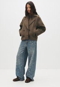 Brown double-breasted wool blend jacket with side pockets, paired with wide-leg blue denim jeans featuring a faded, speckled pattern. Black shoes.