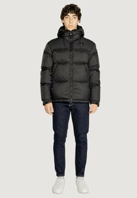 Giubbotto puffer nero con cappuccio, dotato di zip frontale, texture trapuntata e tasche laterali, abbinato a jeans scuri e sneakers bianche.