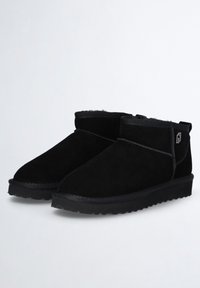 Un par de botas de tobillo de ante negro con forro suave y suelas de goma con relieve, posicionadas una al lado de la otra sobre un fondo simple.
