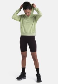 Sudadera verde claro con un bolsillo frontal, combinada con pantalones cortos de bicicleta negros y zapatos de senderismo negros, con puños acanalados y un corte relajado.