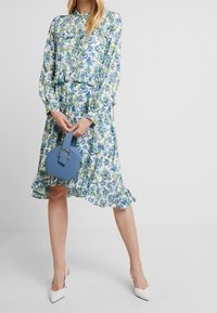 Florale shirtjurk met blauwe en groene patronen, lange mouwen, een cinched taille, gerimpelde zoom, gecombineerd met een ronde blauwe handtas en witte hakken.
