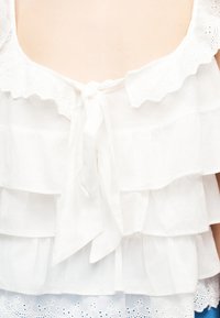 Blouse blanche sans manches avec volants superposés, bordure en dentelle à œillets et lien à nouer à l'encolure.