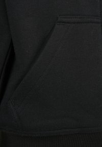Gros plan d'une poche kangourou en tissu noir avec des coutures visibles et un ourlet côtelé, généralement trouvée sur des sweats à capuche ou des sweatshirts.