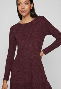 Robe bordeaux, à manches longues côtelées avec un col rond et un corsage ajusté, s'évasant en une jupe froncée. Matériau en tricot texturé.