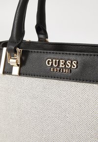 Sac à main Guess texturé noir et blanc avec poignées noires, accessoires dorés, et logo "GUESS EST 1981" sur la garniture en cuir noir.
