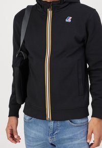 Uomo che indossa una felpa nera con zip, dotata di una cerniera multicolore e del logo K-Way, abbinata a jeans blu e a una borsa a spalla nera.